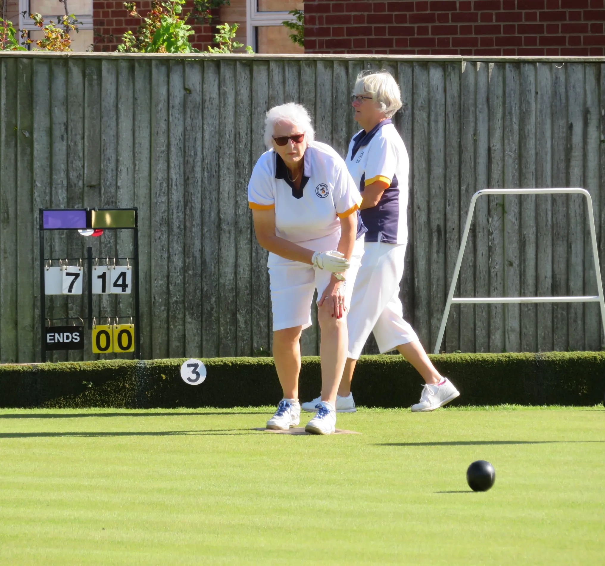 Two Midhurst members in club whites playing on the green, with the rink scoreboard behind them