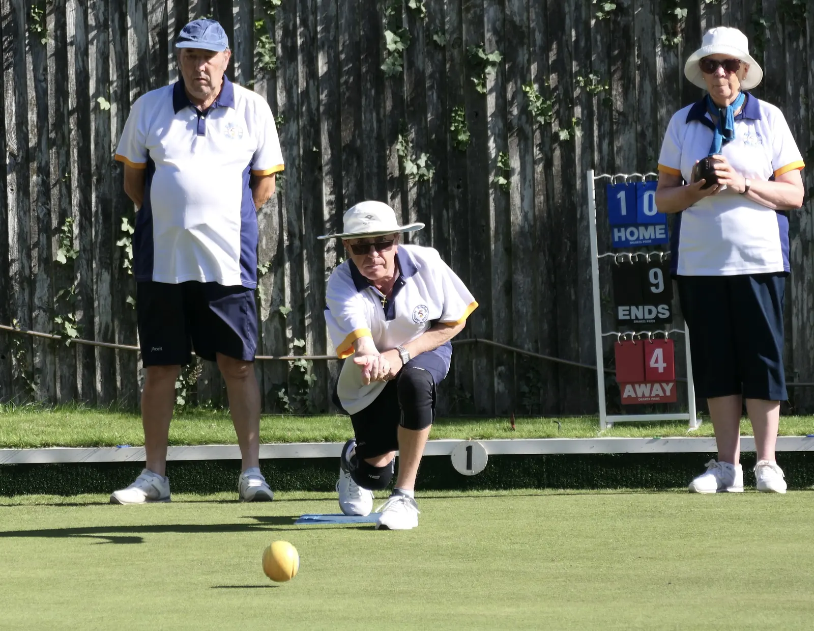 Three Midhurst members in club whites on the green during finals weekend, one of them delivering a bowl, with the rink scoreboard behind them
